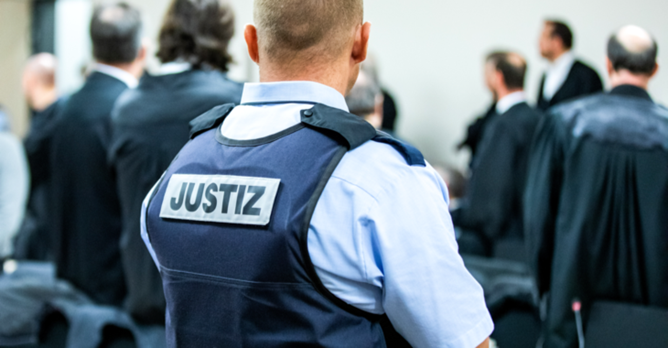 Ein Justizbeamter in einem Gerichtssaal trägt eine Schutzweste mit der Aufschrift Justiz © dpa/Marcel Kusch