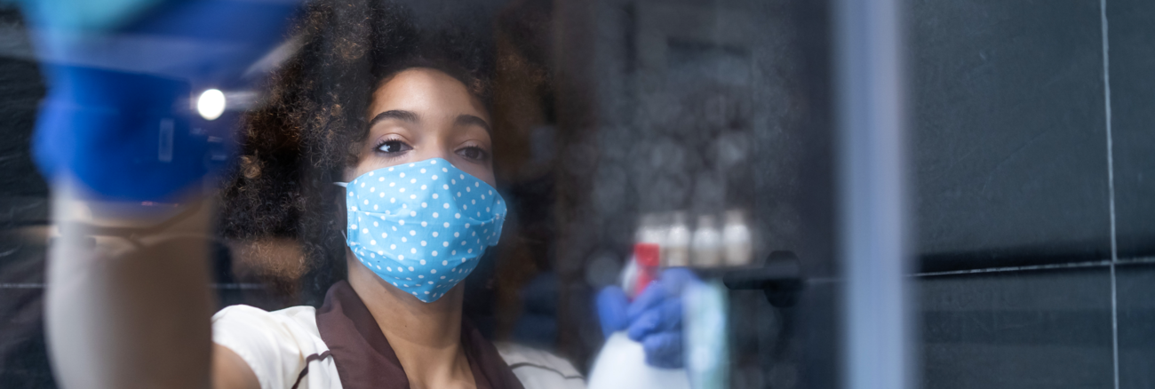 Eine Putzfrau mit Maske, Gummihandschuhen und einer Sprühflasche in der Hand reinigt einen Spiegel © iStock/valentinrussanov Eine Putzfrau mit Maske, Gummihandschuhen und einer Sprühflasche in der Hand reinigt einen Spiegel © iStock/valentinrussanov