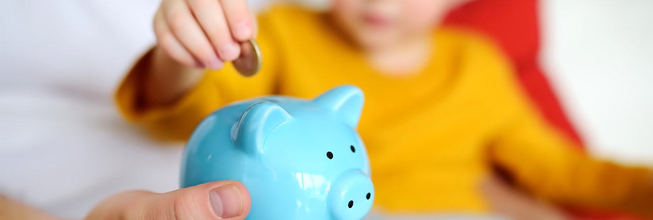 Junge mit gelbem Pullover steckt Euro-Münze in ein hellblaues Sparschwein © iStock/Finanzwissen