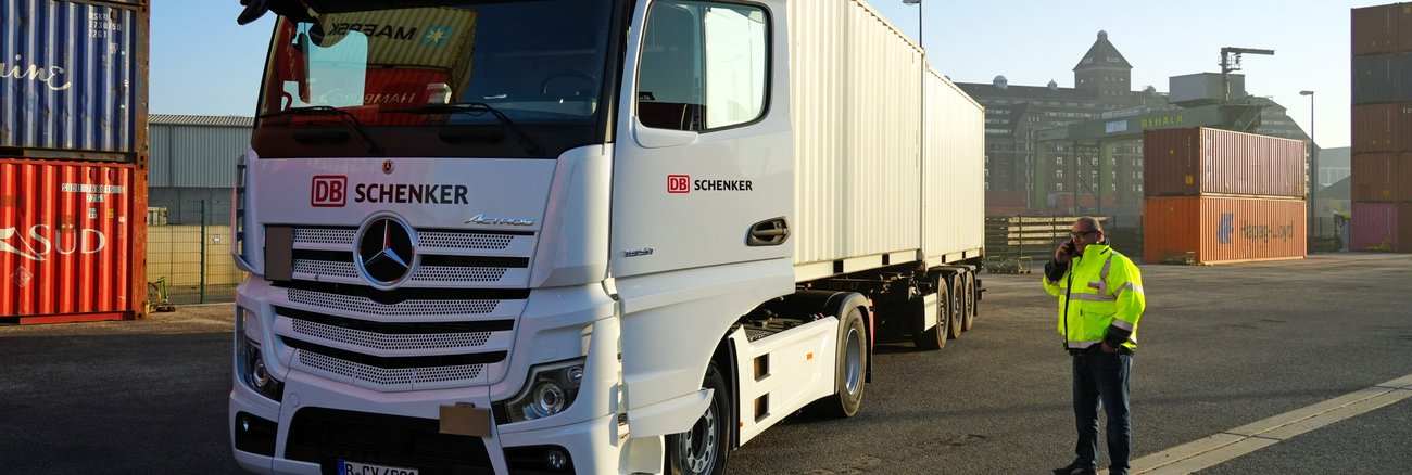 Ein Mann mit gelber Warnweste telefoniert zwischen einem Containern und einem Lkw mit der Aufschrift DB Schenker © Deutsche Bahn AG/Volker Emersleben