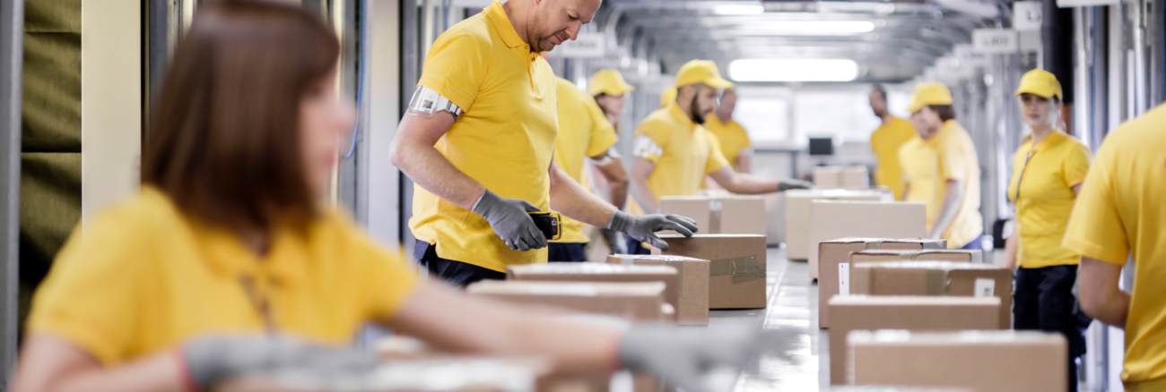 Arbeiter:innen in einem Logistikunternehmen verpacken an einem Fließband Pakete @ iStock/simonkr Arbeiter:innen in einem Logistikunternehmen verpacken an einem Fließband Pakete @ iStock/simonkr