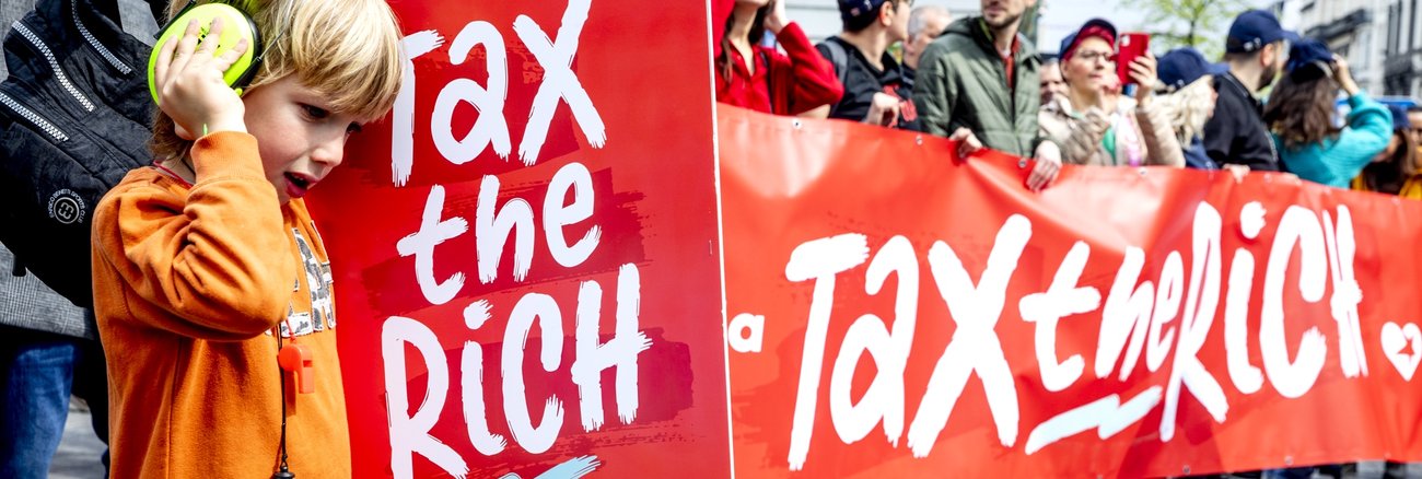 Teilnehmer an einer Demonstration halten ein Transparent und ein Plakat mit der Aufschrift: Tax the rich © picture alliance/dpa/Belga|Hatim Kaghat Teilnehmer an einer Demonstration halten ein Transparent und ein Plakat mit der Aufschrift: Tax the rich © picture alliance/dpa/Belga|Hatim Kaghat