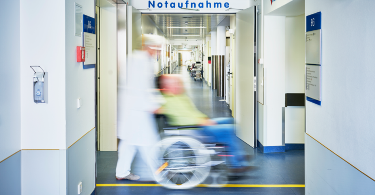Ein Pfleger schiebt in einem Krankenhaus einen Patienten im Rollstuhl durch einen Flur mit einem Schild Notaufnahme an der Wand © iStock/upixa Ein Pfleger schiebt in einem Krankenhaus einen Patienten im Rollstuhl durch einen Flur mit einem Schild Notaufnahme an der Wand © iStock/upixa