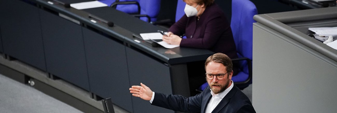 Jan Korte spricht im Plenum mit Angela Merkel im Hintergrund