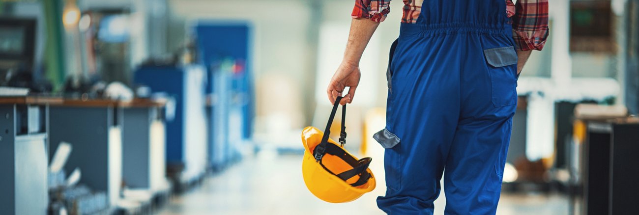 Ein Arbeit in blauer Latzhose und mit gelbem Helm in der Hand läuft durch eine Werkhalle © iStock/gilaxia