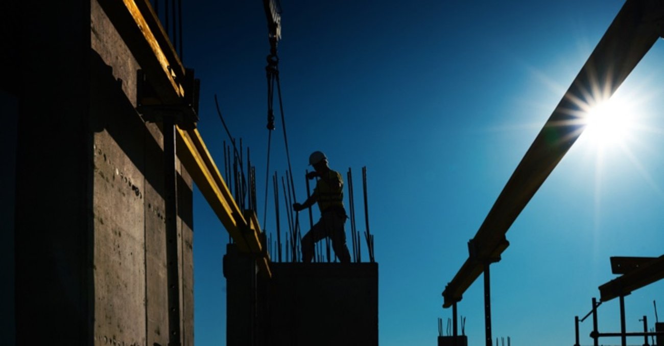 Silhouette eines Bauarbeiters auf einer Baustelle mit Stahlträgern, Kran und Betonpfeilern © iStock/gilaxia