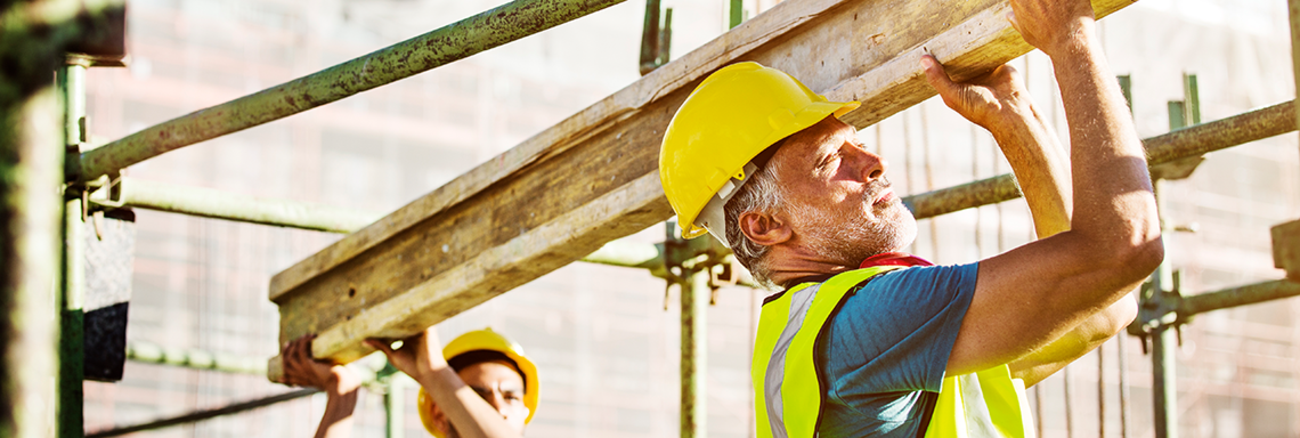 Zwei Arbeiter mit Helm und Schutzweste stemmen einen Stahlträger auf einer Baustelle. Foto: © istock.com/Morsa Images Zwei Arbeiter mit Helm und Schutzweste stemmen einen Stahlträger auf einer Baustelle. Foto: © istock.com/Morsa Images