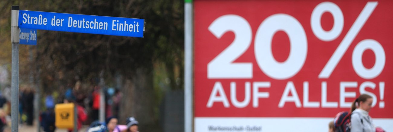 Passanten laufen in Wolmirstedt (Sachsen-Anhalt) an einem Straßenschild mit der Aufschrift »Straße der deutschen Einheit« vorbei © picture alliance/dpa|Jens Wolf