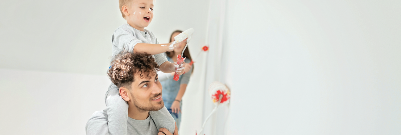 Eine Junge Familie beim Malern ihrer Wohnung, der Sohn mit Farbrolle auf den Schultern des Vaters © iStock/aywan88