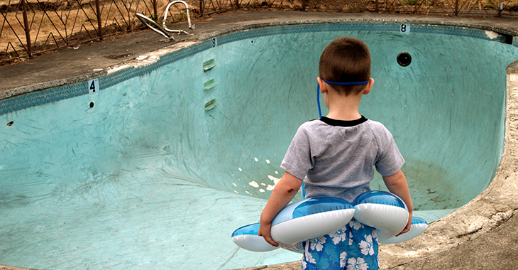 Ein kleiner Junge mit Schwimmhilfe steht vor einem leeren, renovierungsbedürftigen Schwimmbecken in einem Freibad. Foto: © iStock.com/RickLordPhotography Ein kleiner Junge mit Schwimmhilfe steht vor einem leeren, renovierungsbedürftigen Schwimmbecken in einem Freibad. Foto: © iStock.com/RickLordPhotography
