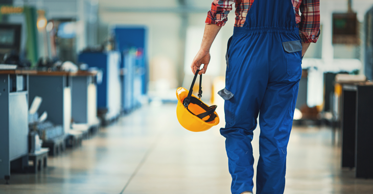 Ein Arbeit in blauer Latzhose und mit gelbem Helm in der Hand läuft durch eine Werkhalle © iStock/gilaxia Ein Arbeit in blauer Latzhose und mit gelbem Helm in der Hand läuft durch eine Werkhalle © iStock/gilaxia