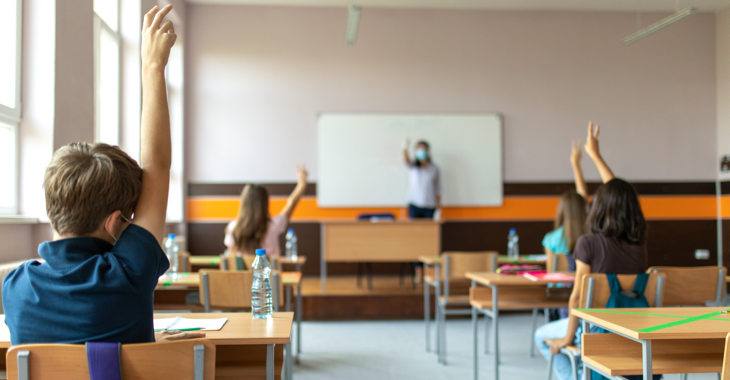 Schüler:innen mit Masken in einem Klassenzimmer melden sich, im Hintergrund eine Lehrerin mit Maske vor der Tafel © iStock/miljko